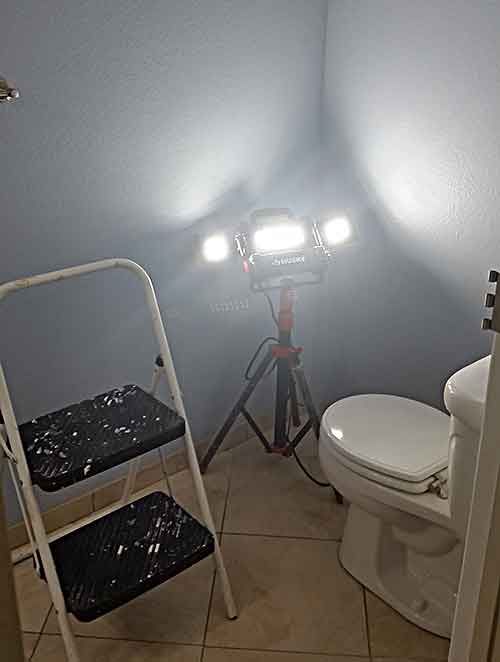 This image shows the light having its tripod partially collapsed against a bathroom corner and with the light stand fully drawn down so the light is as low to the floor as possible. The light is turned on and you can see the light pattern of the two side panels brighten two walls.