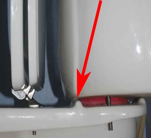 The image shows a side view and very close view of the toilet seat on the left in a up osition. In the center of the image shows the first tank ridge which the water tank rests upon. A red arrow points to where the shim was inserted before the tank was tightened. The bolt for mounting the tank can be seen. Near the right edge of the image is the rear tank mounting ridge.